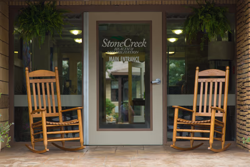 Two wooden rocking chairs on a tiled porch in front of a glass door with the text 'StoneCreek Health & Rehabilitation MAIN ENTRANCE'. The door is framed by brick walls and there are green plants hanging above the chairs.