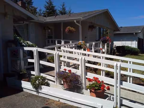 Front exterior of a senior living home with white railings and potted flowers along a walkway.