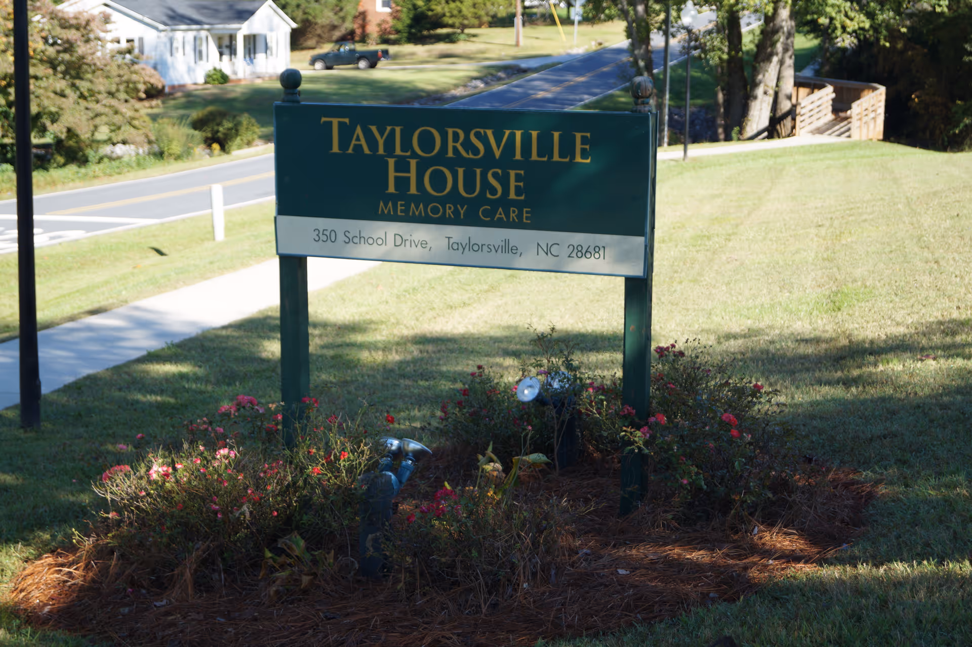 Green sign on a lawn reading "Taylorsville House Memory Care" with the address and surrounding landscaping and a road in the background.