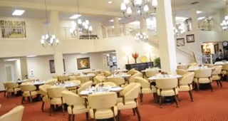 A spacious dining room with multiple round tables surrounded by beige upholstered chairs. The room features red carpet flooring, cream-colored walls, and several chandeliers hanging from the ceiling. There is a staircase and a balcony area visible in the background, along with framed artwork on the walls.
