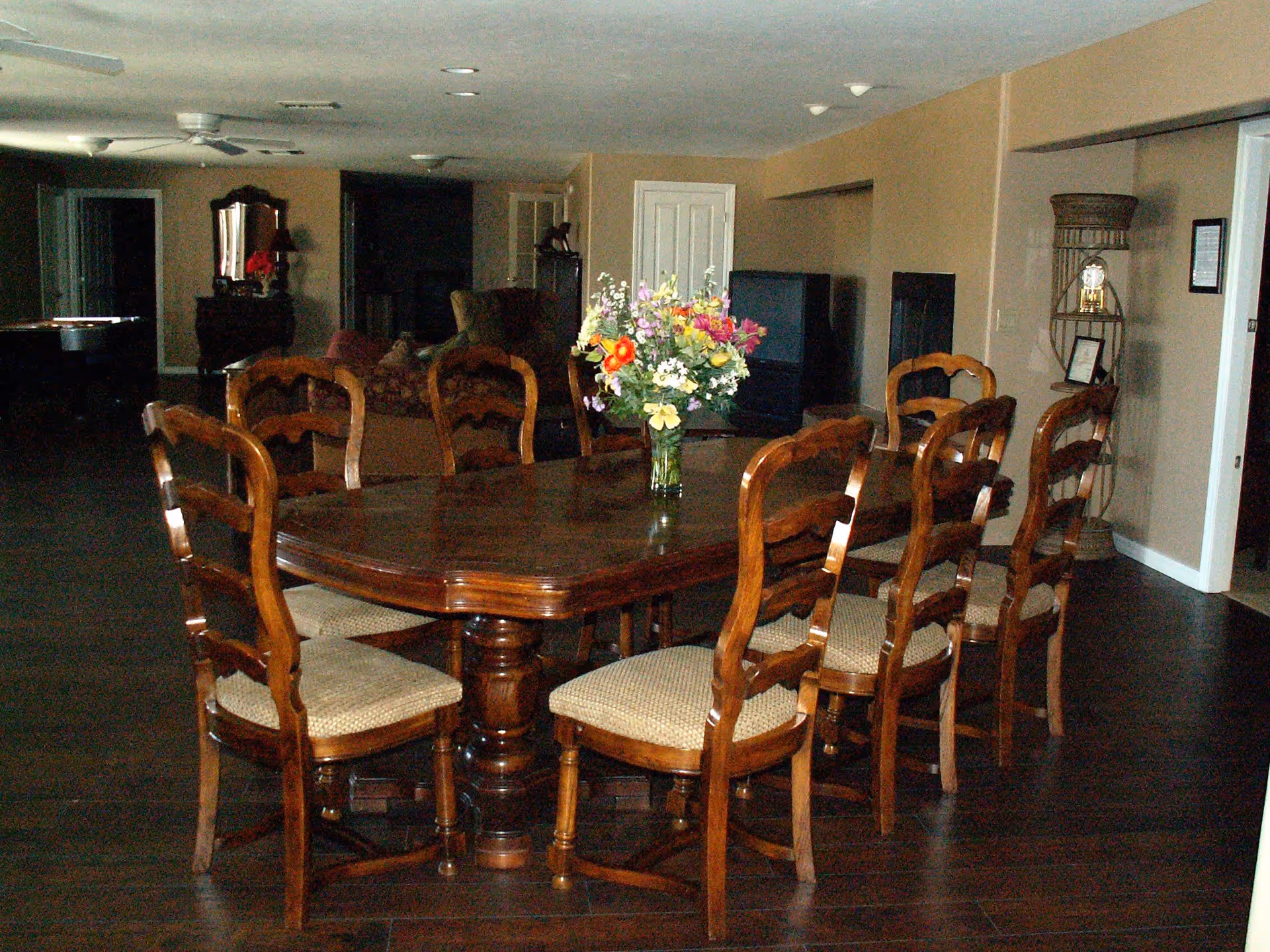 A dining room with a large wooden table surrounded by eight wooden chairs with cushioned seats. A vase with colorful flowers is placed in the center of the table. In the background, there is a living area with sofas, a TV, and a decorative shelf with a clock and framed pictures. The room has dark wooden flooring and beige walls.