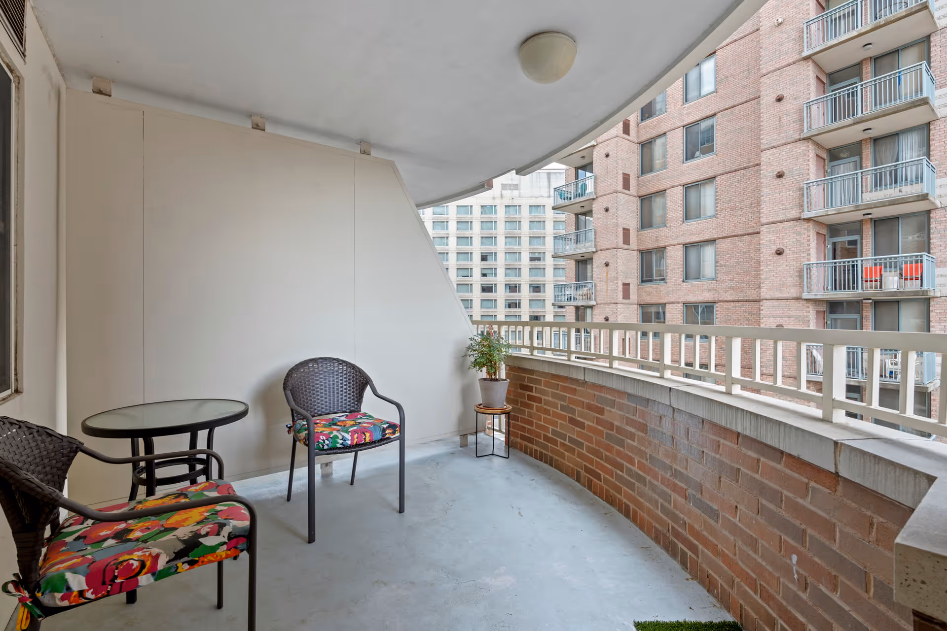 A curved balcony with a brick half-wall railing, two wicker chairs with colorful floral cushions, a small round glass-top table, and a potted plant on a small stand. The balcony overlooks nearby apartment buildings.