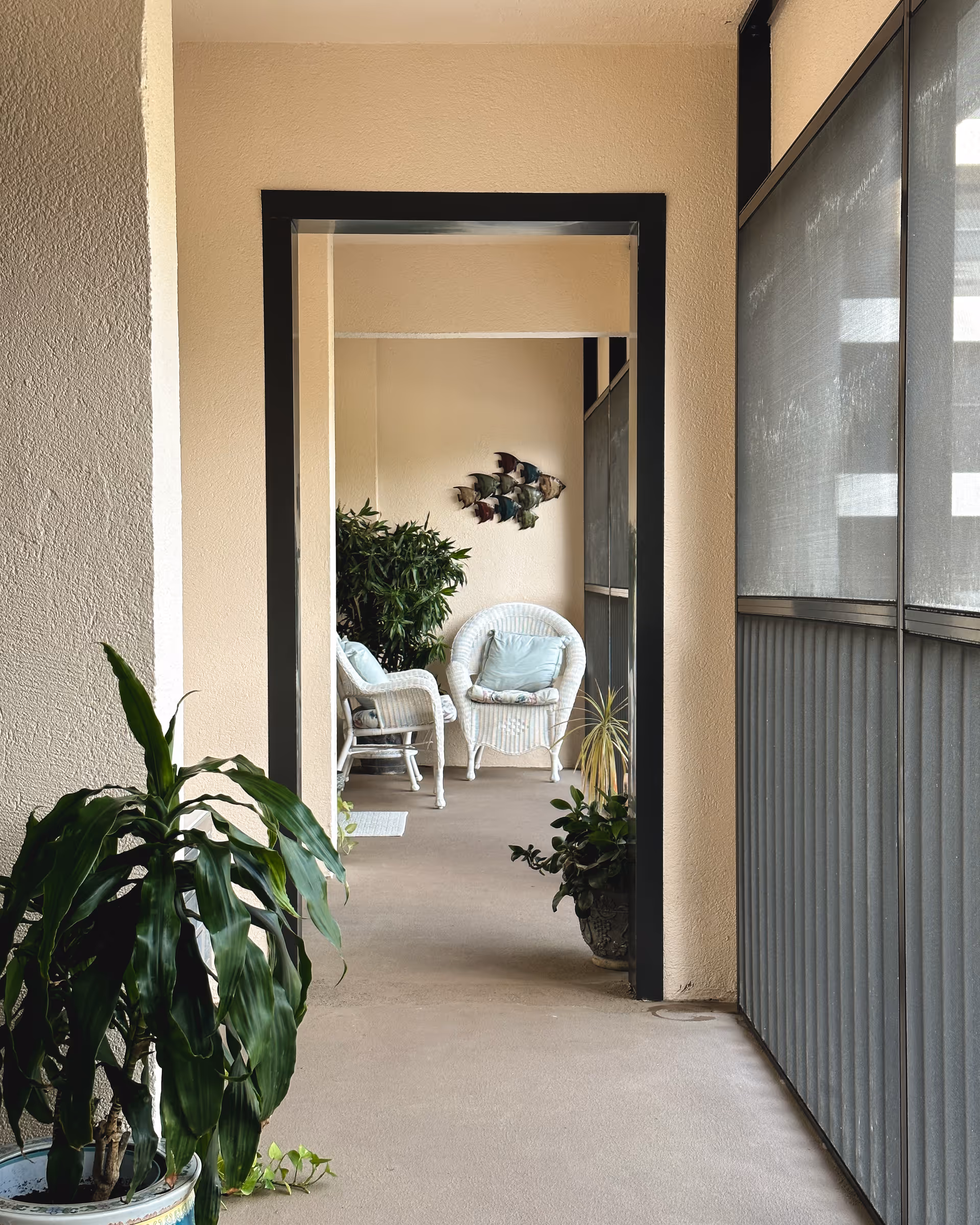 A screened porch corridor featuring white wicker chairs, multiple potted plants, and decorative fish wall art.