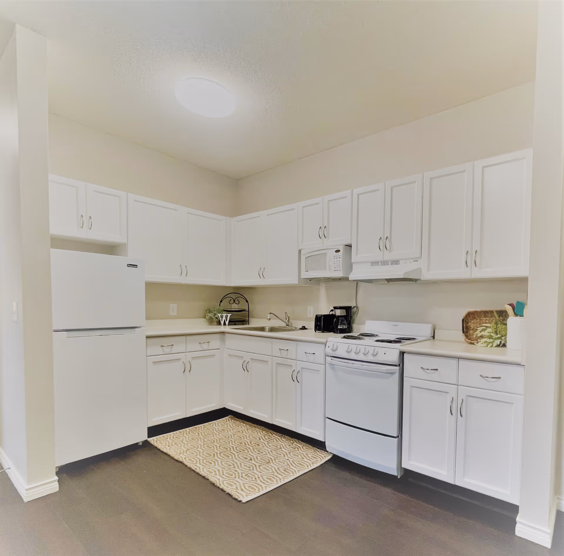 A clean, modern kitchen with white cabinets, a white refrigerator, a white stove with an oven, a microwave above the stove, a coffee maker on the counter, and a small patterned rug on the dark floor.