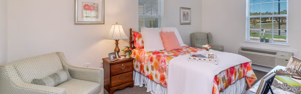 A bright and cozy bedroom featuring a bed with a colorful floral bedspread and pink pillows. Next to the bed is a wooden nightstand with a lamp, a framed photo, and a small vase of flowers. There is a cushioned armchair in the corner near a window with blinds, and another patterned armchair with a pillow on the opposite side. The room has light-colored walls with framed artwork and a window air conditioning unit beneath one window.
