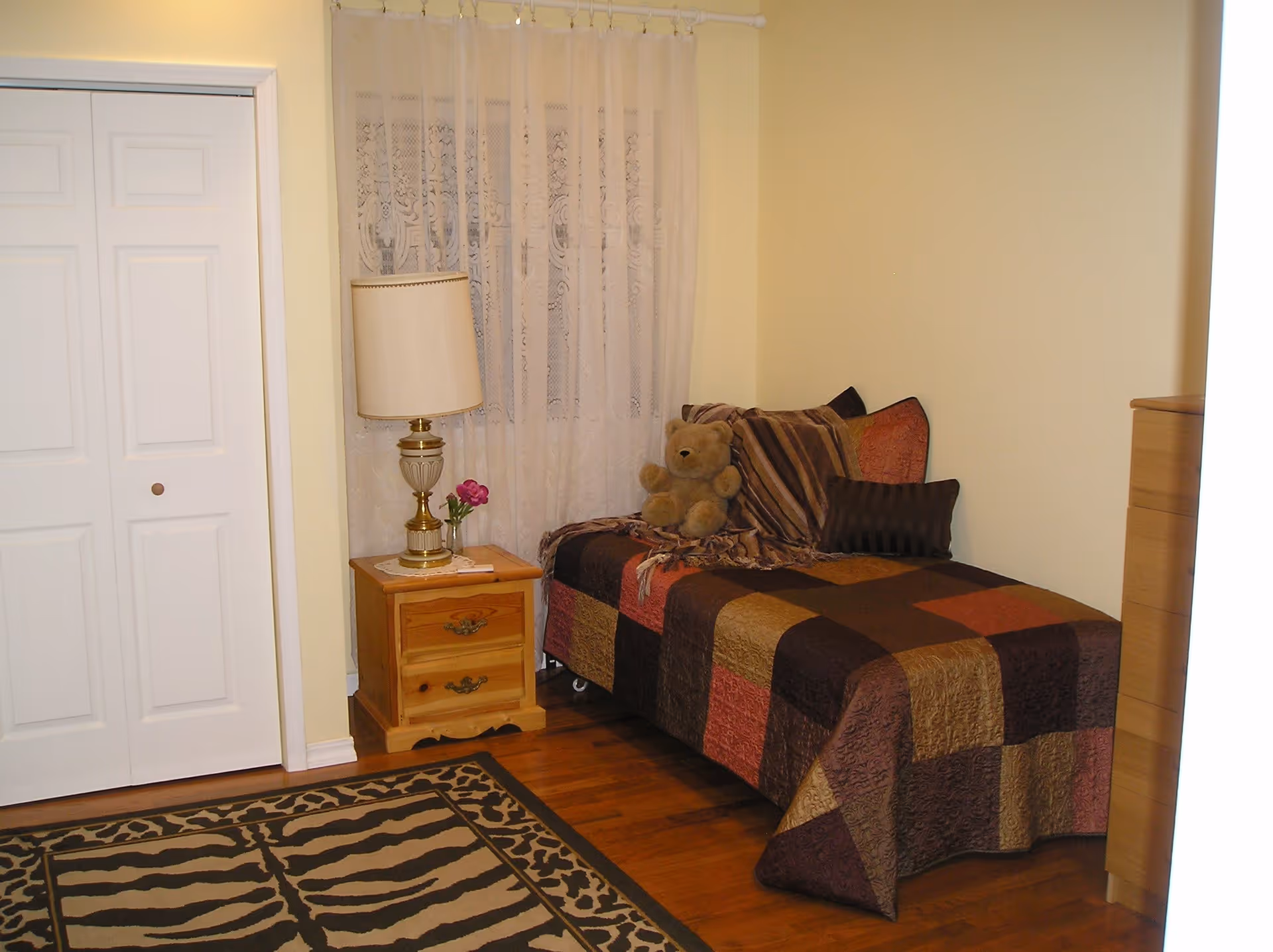 A cozy bedroom with a single bed covered in a patchwork quilt in shades of brown, orange, and gold. There are several pillows and a teddy bear on the bed. Next to the bed is a wooden nightstand with a decorative lamp and a small vase with a pink flower. A window with sheer lace curtains is behind the nightstand. The floor has a patterned rug with animal print designs. A white closet door is visible on the left side of the room.