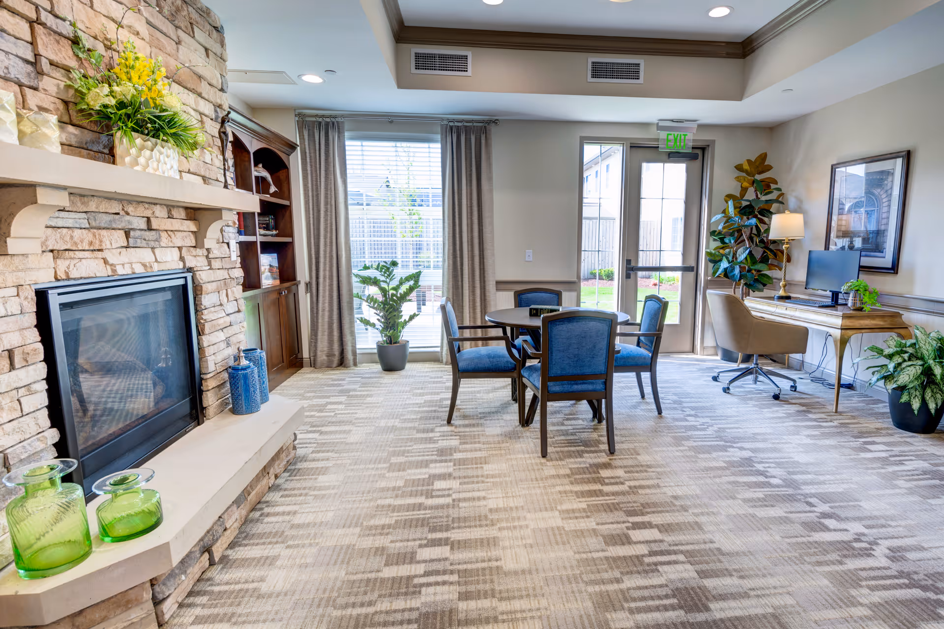 A bright and spacious living room area with a stone fireplace on the left, decorated with green plants and blue vases. In the center, there is a round table with four blue upholstered chairs. To the right, there is a wooden desk with a computer monitor, a lamp, and a beige swivel chair. Large windows with beige curtains and a glass door lead to an outdoor area. The room has a patterned carpet and neutral-colored walls.