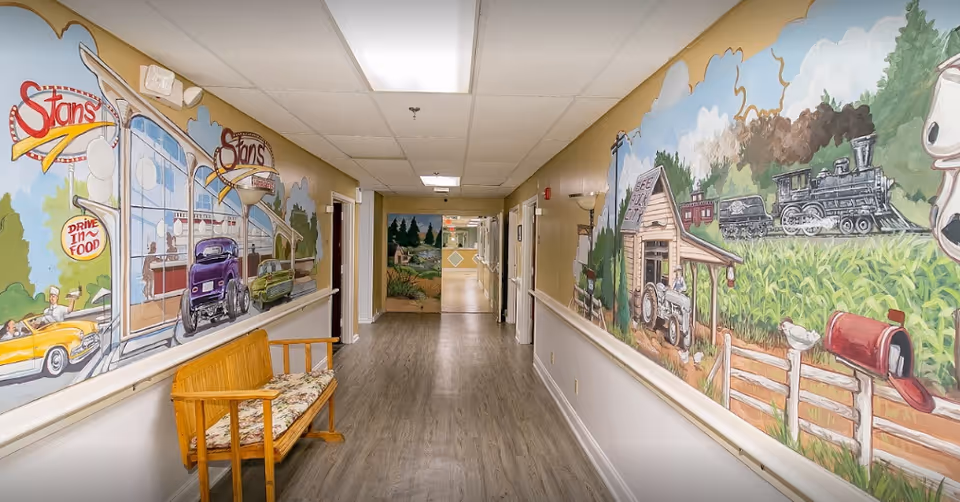 Interior hallway in a senior living facility featuring colorful wall murals of a diner and countryside, a wooden bench, handrails, and wood-look flooring.