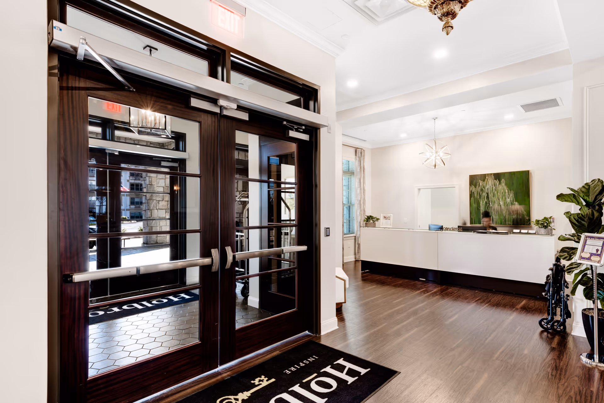 Entrance area of Holbrook Decatur facility showing double glass doors with dark wood frames and metal push bars. Inside, there is a reception desk with a person behind it, a modern chandelier, a large green abstract painting on the wall, and a wheelchair parked near a potted plant. The floor is dark wood and there is a black mat with the Holbrook logo near the entrance.