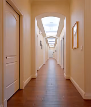 A long, well-lit hallway with wooden flooring and cream-colored walls. There are multiple closed doors on both sides of the hallway and framed pictures hanging on the walls. The ceiling features recessed lighting with arched architectural details.