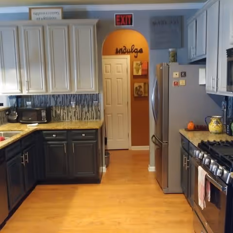 A modern kitchen with light and dark cabinetry, granite countertops, a stainless steel refrigerator, a microwave, and a gas stove. The floor is wooden, and there is an arched doorway with the word 'indulge' above it. Various decorative items and a sign that reads 'Happiness is a journey, not a destination' are visible.