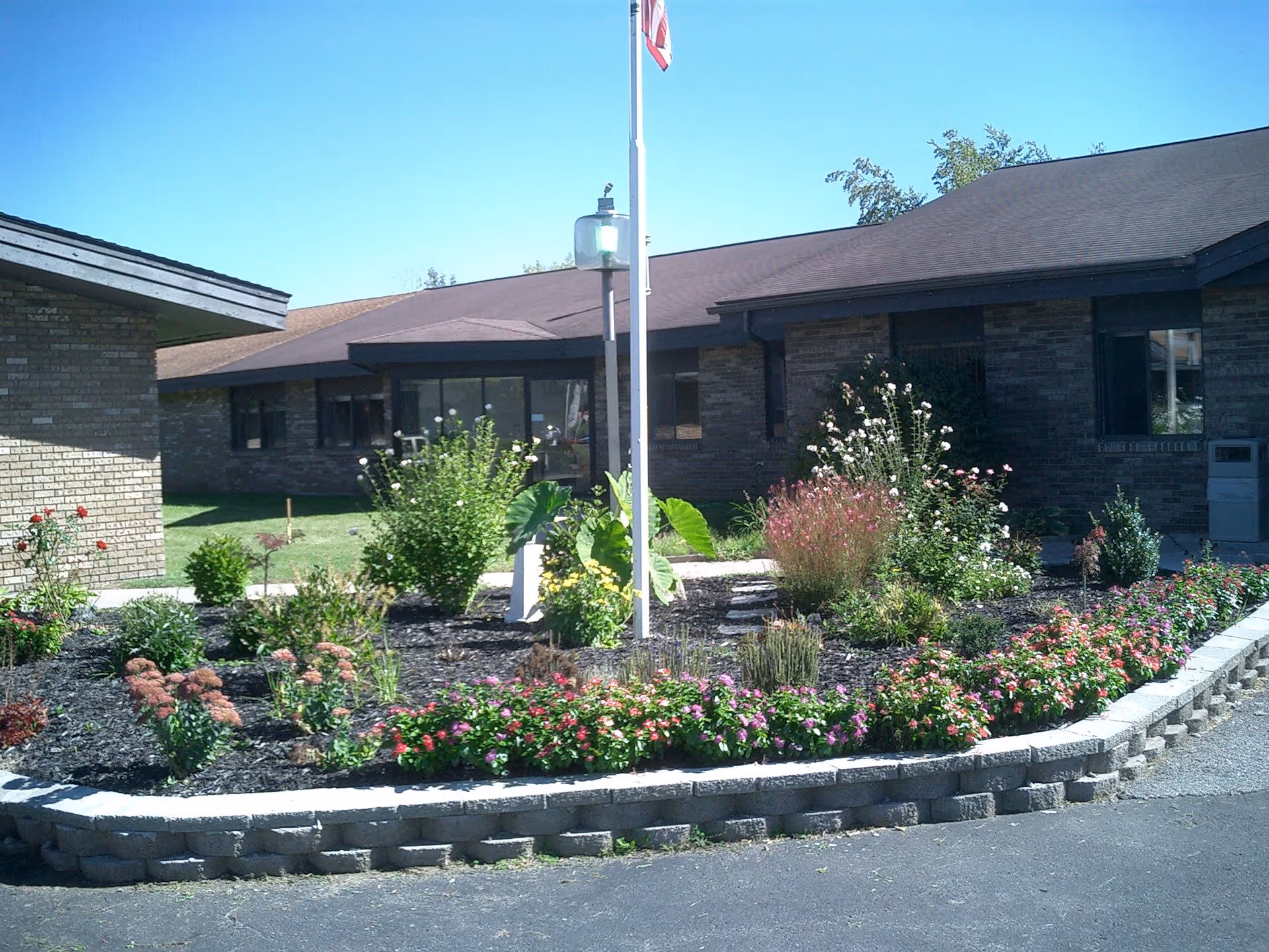 A landscaped garden bed with various flowering plants and shrubs in front of a single-story brick building under a clear blue sky. A flagpole with an American flag is centered in the garden bed.