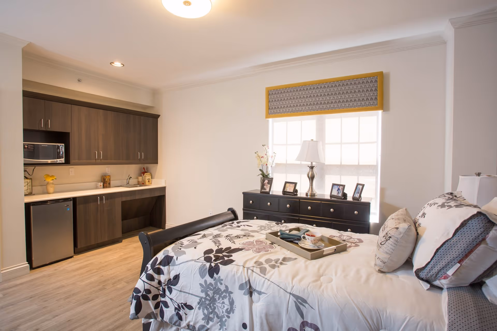 A well-lit bedroom with a bed featuring floral patterned bedding and multiple pillows. A tray with a cup and napkin is placed on the bed. To the left, there is a kitchenette area with dark wood cabinets, a small refrigerator, a microwave, and a countertop with a sink. In front of the bed, a black dresser holds a lamp and several framed photos. A large window with a decorative valance allows natural light into the room.