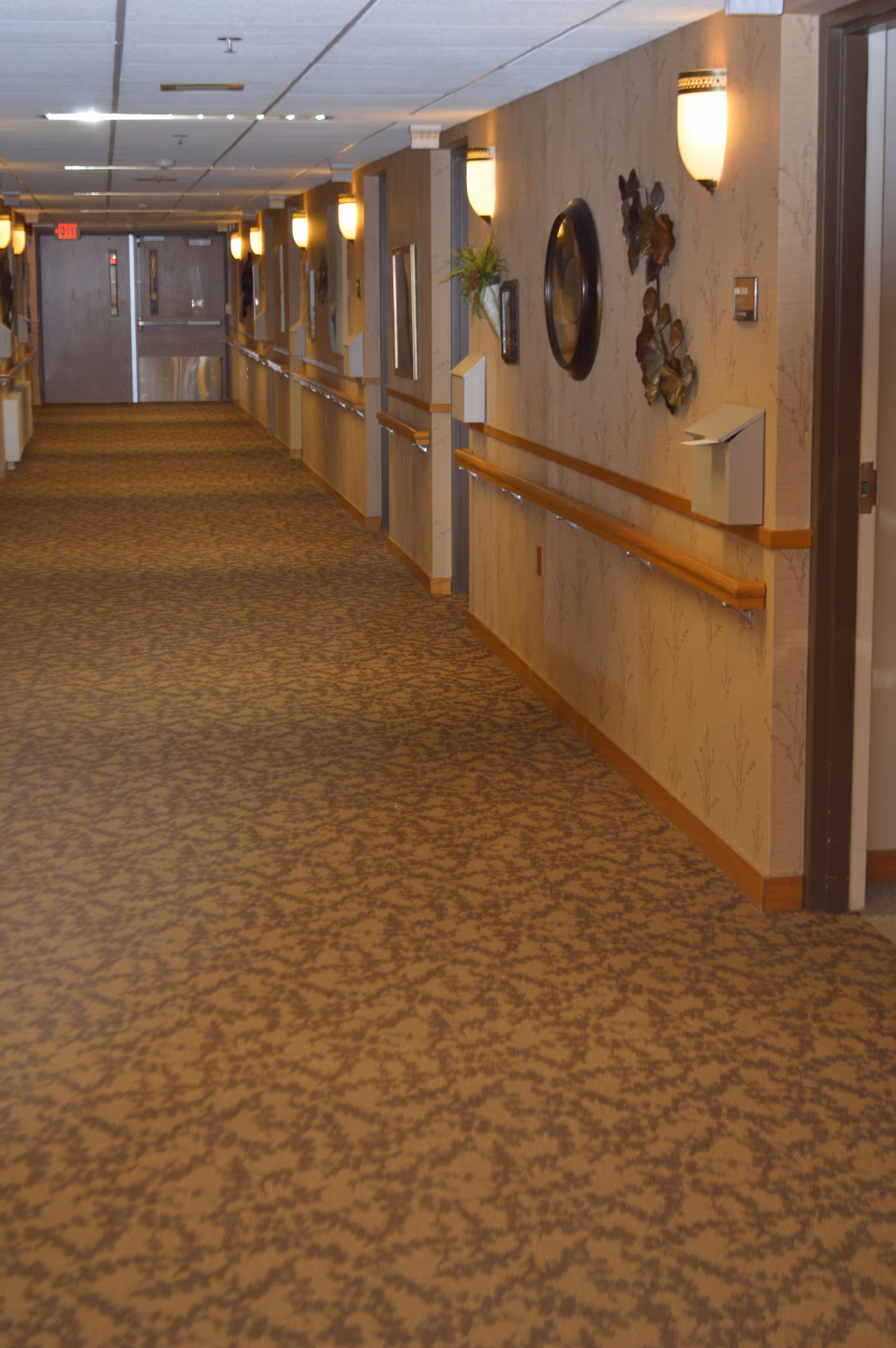 A long, carpeted hallway in an assisted living facility with beige patterned carpet and walls. The walls have wooden handrails and are decorated with framed pictures, a round mirror, and metal wall art. Wall-mounted lights illuminate the corridor, and double doors are visible at the far end.