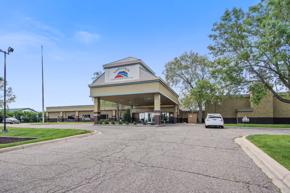 Front exterior of a single-story senior living facility with a covered entrance/porte-cochere, parking lot, and surrounding trees under a blue sky.