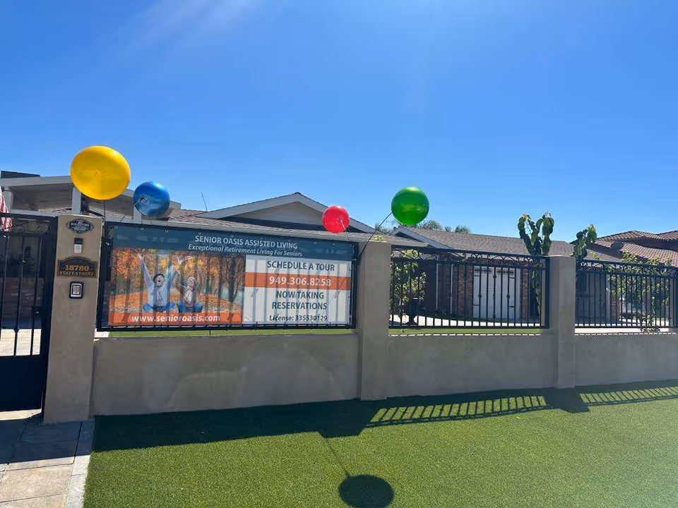 Exterior view of Senior Oasis Assisted Living facility with a beige wall and black metal fence. There are colorful balloons (yellow, blue, red, green) attached to the fence. A large banner on the fence advertises Senior Oasis Assisted Living, showing two seniors enjoying outdoors and providing a phone number and website for scheduling a tour. The sky is clear and blue.