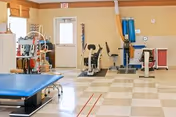 A rehabilitation/exercise room with therapy tables, exercise bikes, and medical equipment on a tiled floor near a door.