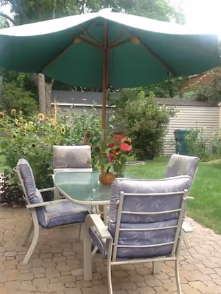 Outdoor patio area with a glass-top table surrounded by four cushioned chairs. A large green umbrella shades the table. There are flowers in a vase on the table and greenery including bushes and trees in the background.