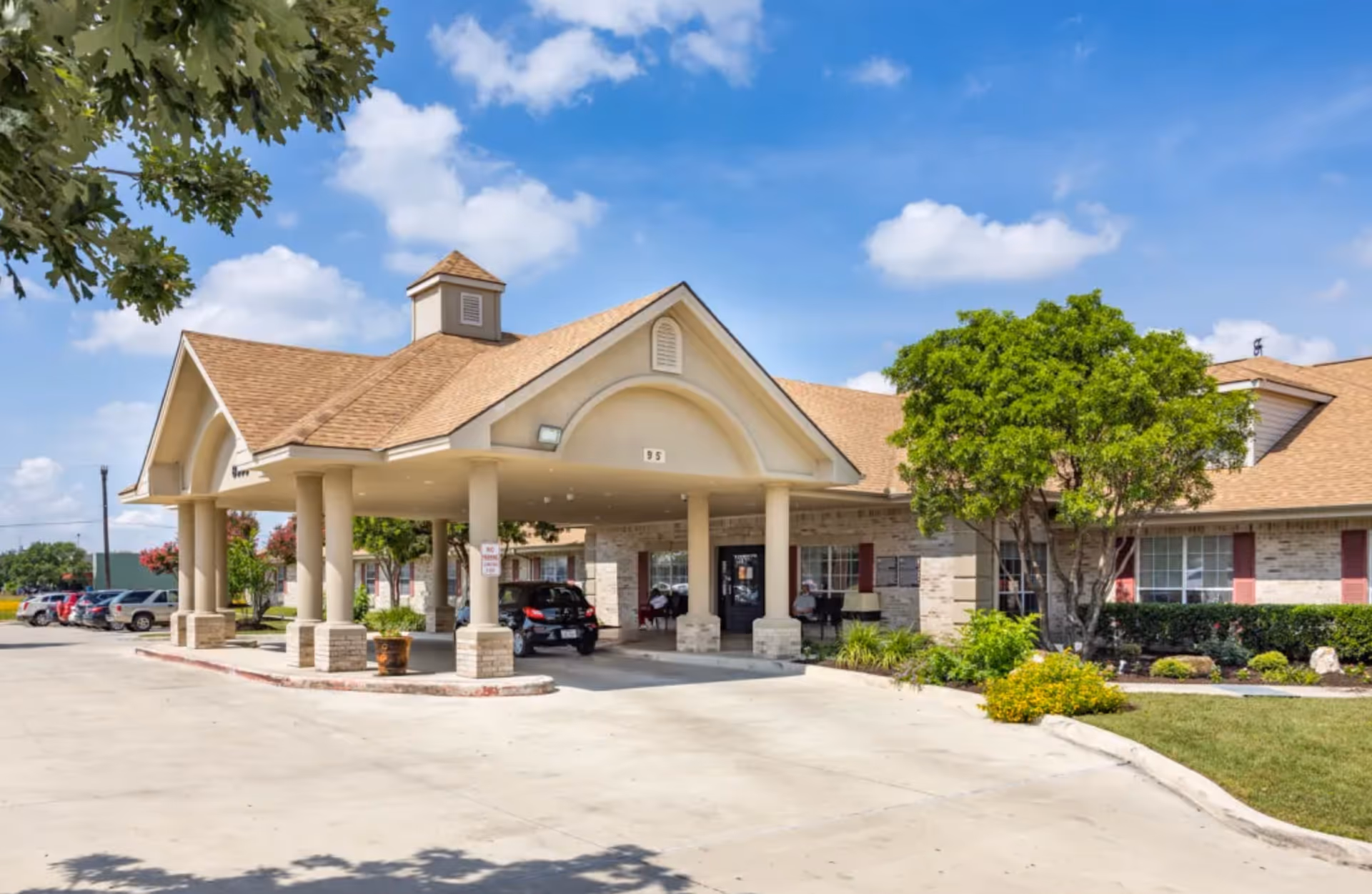 Exterior view of Mystic Park Nursing and Rehabilitation Center showing the main entrance with a covered drop-off area supported by columns, surrounded by landscaped greenery and a parking lot with several cars under a partly cloudy blue sky.