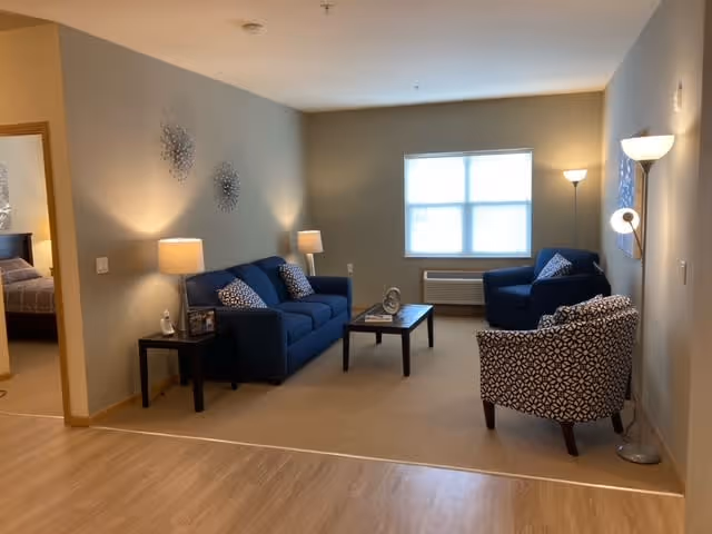 A cozy senior living room with a blue sofa, two patterned armchairs, a coffee table, and two side tables with lamps. The room has beige walls and carpet, a large window letting in natural light, and a floor lamp in the corner. A bedroom is visible through an open doorway on the left.
