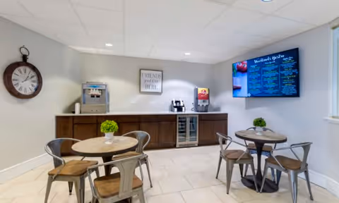 A small dining area with two round wooden tables, each surrounded by four metal chairs with wooden seats. On each table is a small green plant centerpiece. Against the back wall is a wooden cabinet with a beverage dispenser, coffee machine, and a small refrigerator. A large digital menu board is mounted on the right wall, and a round wall clock hangs on the left wall. The room has light-colored tiled flooring and a white ceiling with recessed lighting.