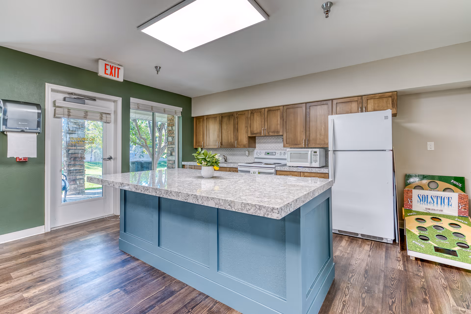 A bright kitchen area in a senior living facility featuring a large island with a marble countertop and blue base. The kitchen has wooden cabinets, a white refrigerator, microwave, and stove. There is a door and window with a view of greenery outside. A game board with the text 'SOLSTICE' is leaning against the wall near the refrigerator. The walls are painted green and beige, and the floor is wood.