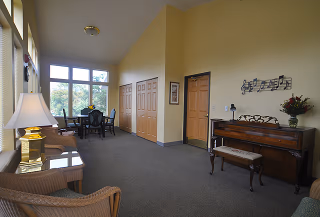 Bright communal lounge with wicker seating, a piano, and a dining table by large windows.