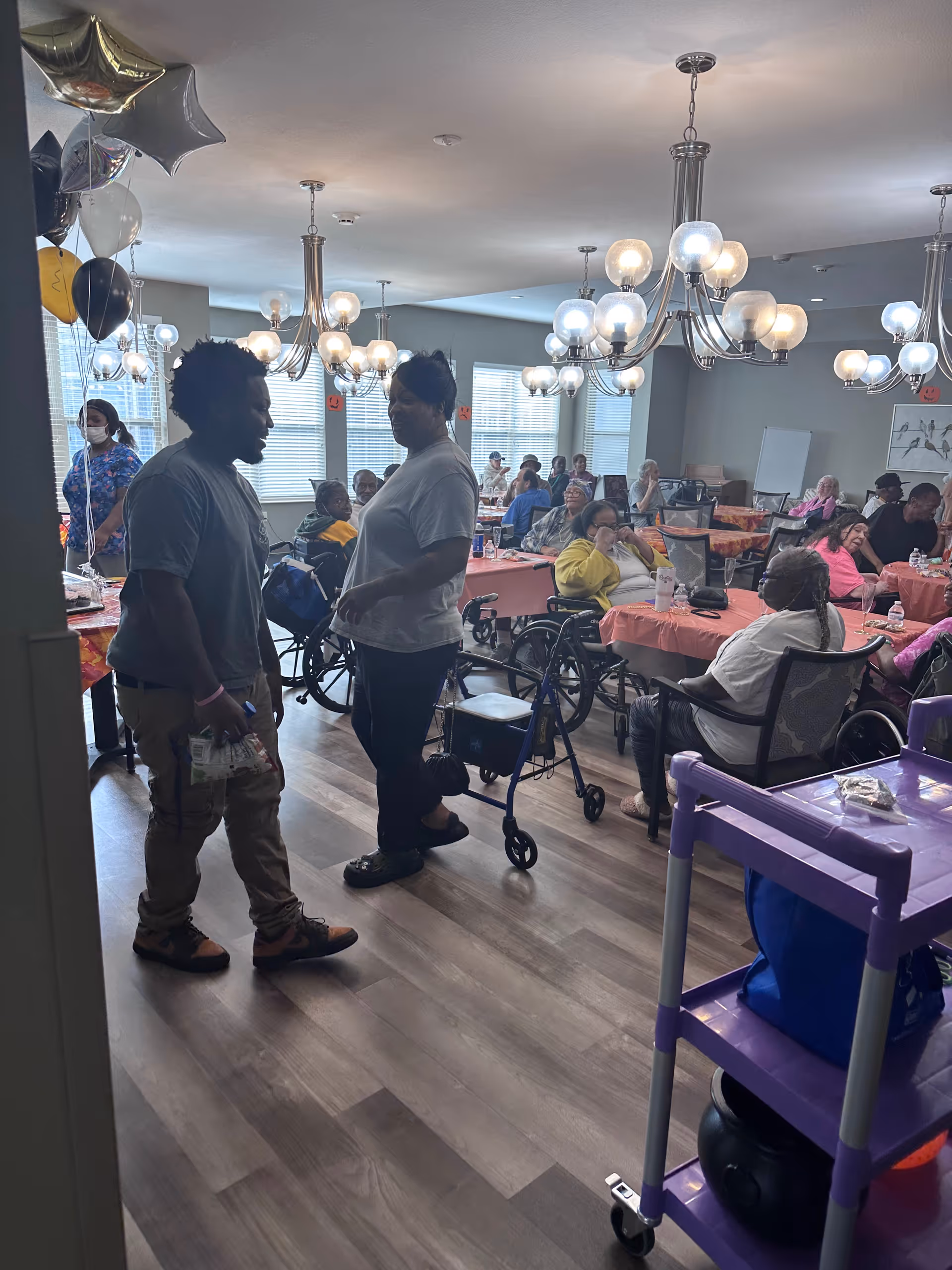 Residents and staff gathered in a decorated common dining/activity room with tables, wheelchairs, and chandeliers.