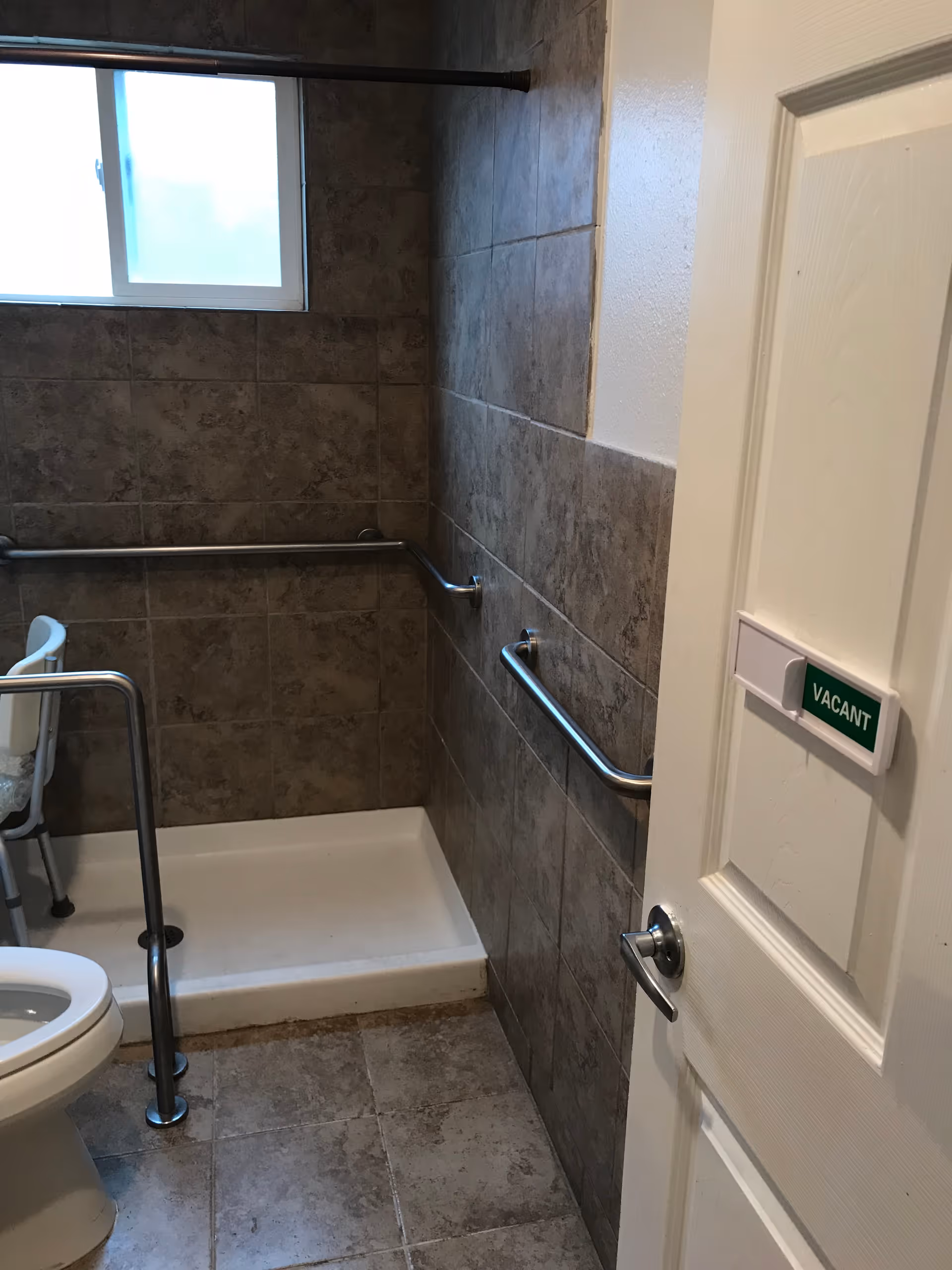 Accessible bathroom with a toilet, walk-in shower, multiple grab bars, a shower chair and a 'VACANT' sign on the door.