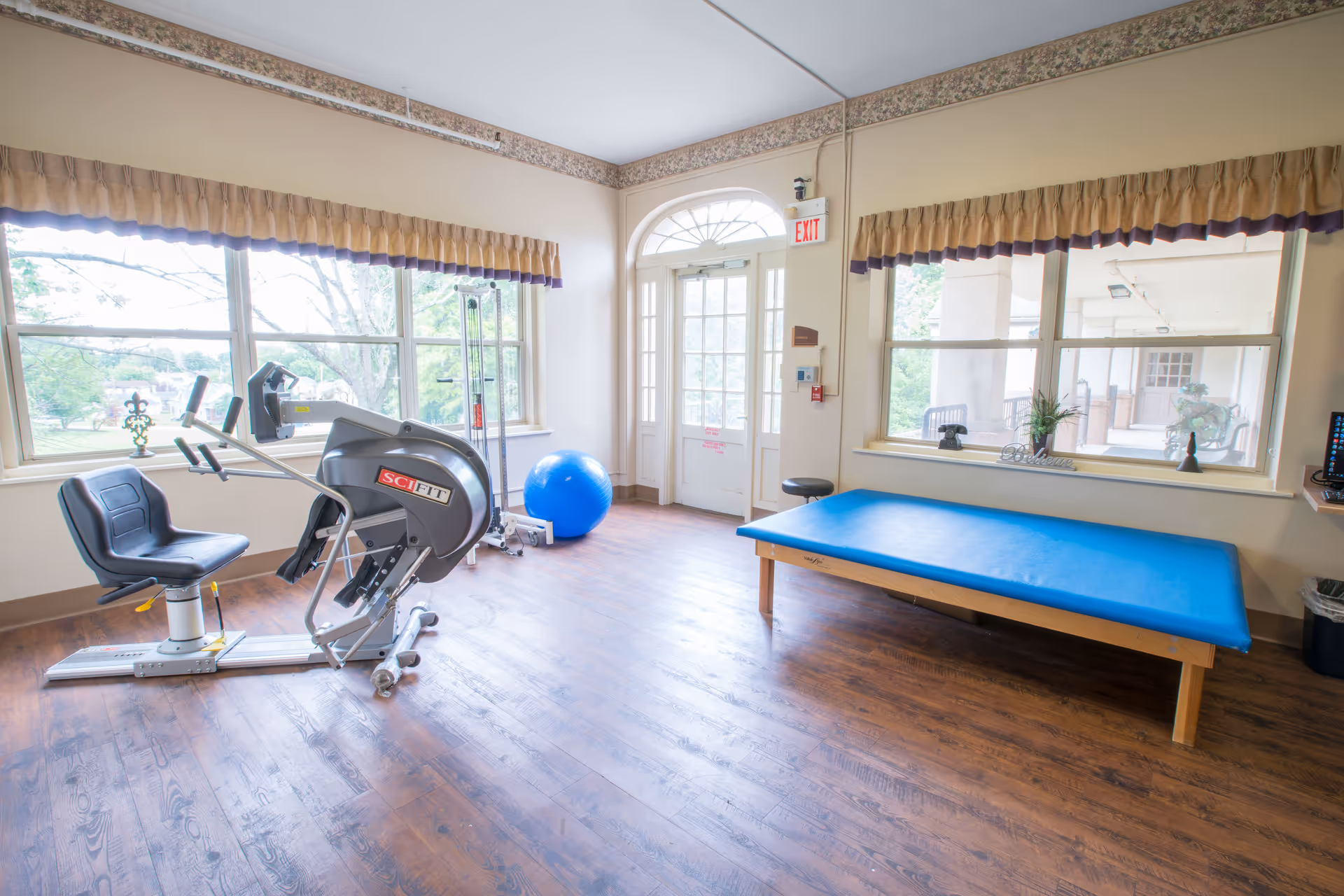 A bright exercise room with large windows, a recumbent exercise bike, a blue exercise ball, and a blue padded therapy table. The room has wooden flooring, beige walls with a decorative border near the ceiling, and an exit door with a window above it. Outside the windows, greenery and a covered patio area are visible.