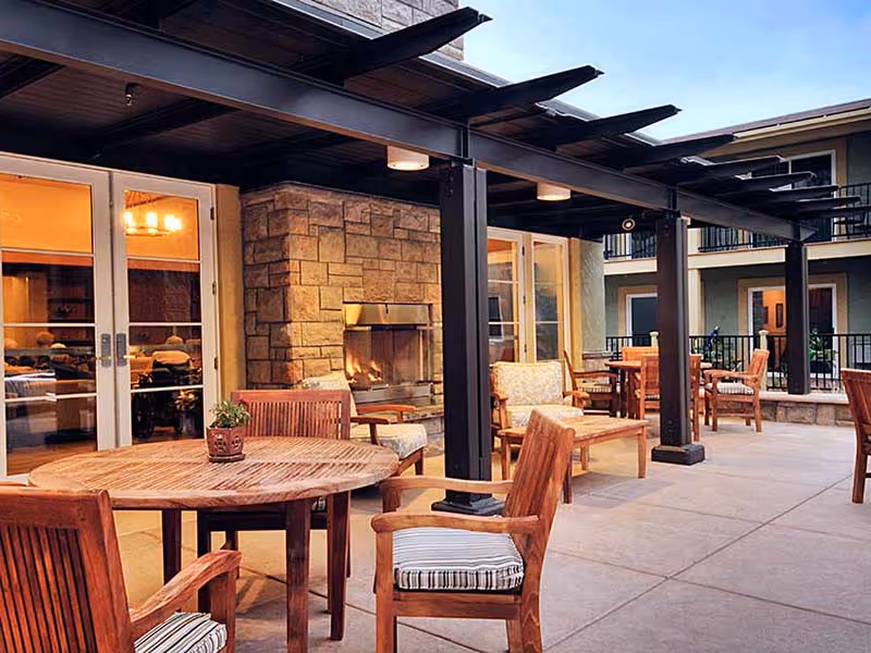 Outdoor patio area with wooden tables and chairs, a stone fireplace, and a covered pergola structure attached to a building with glass doors leading inside.