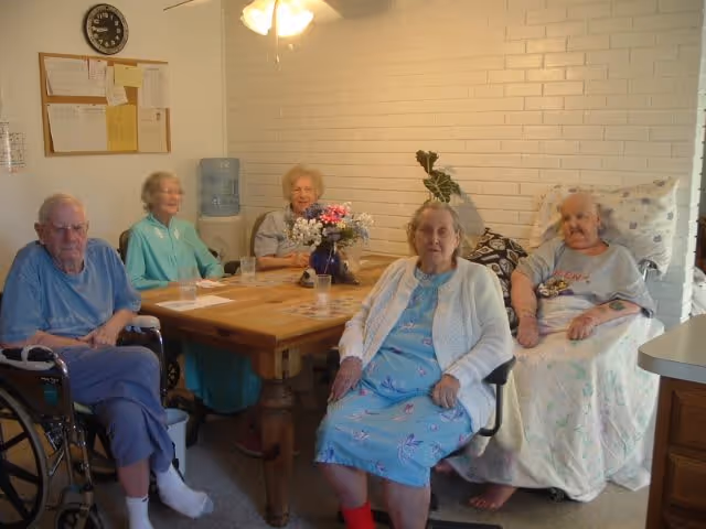 Five elderly individuals sitting around a wooden table in a room with white brick walls. One person is in a wheelchair, two are seated on chairs, and two are sitting on beds. There is a vase with flowers on the table and a water dispenser in the background.