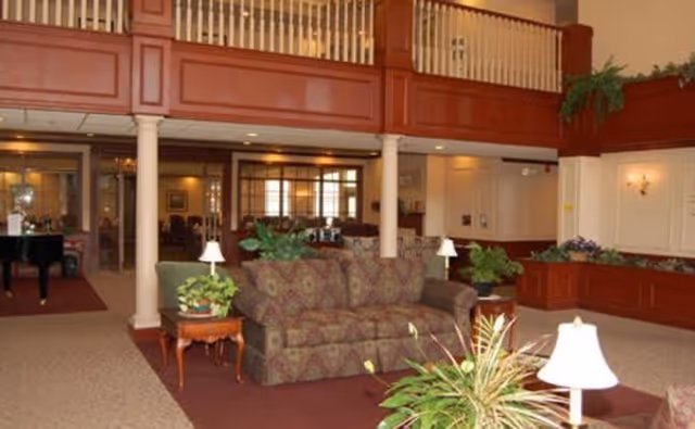 A spacious living room area in Brentwood at Elkhart Independent Living featuring a patterned sofa, side tables with lamps, several potted plants, and a grand piano in the background. The room has a high ceiling with a balcony railing above and warm wood paneling accents.