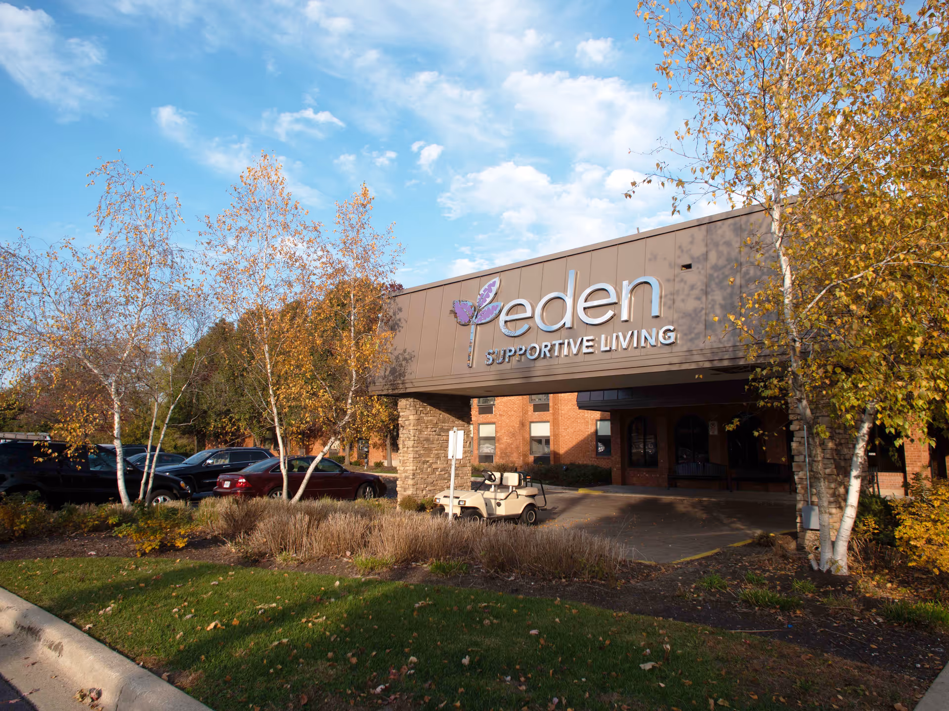 Exterior view of Eden Supportive Living Fox Valley building entrance with a covered drop-off area, surrounded by trees with autumn foliage and parked cars nearby under a partly cloudy sky.