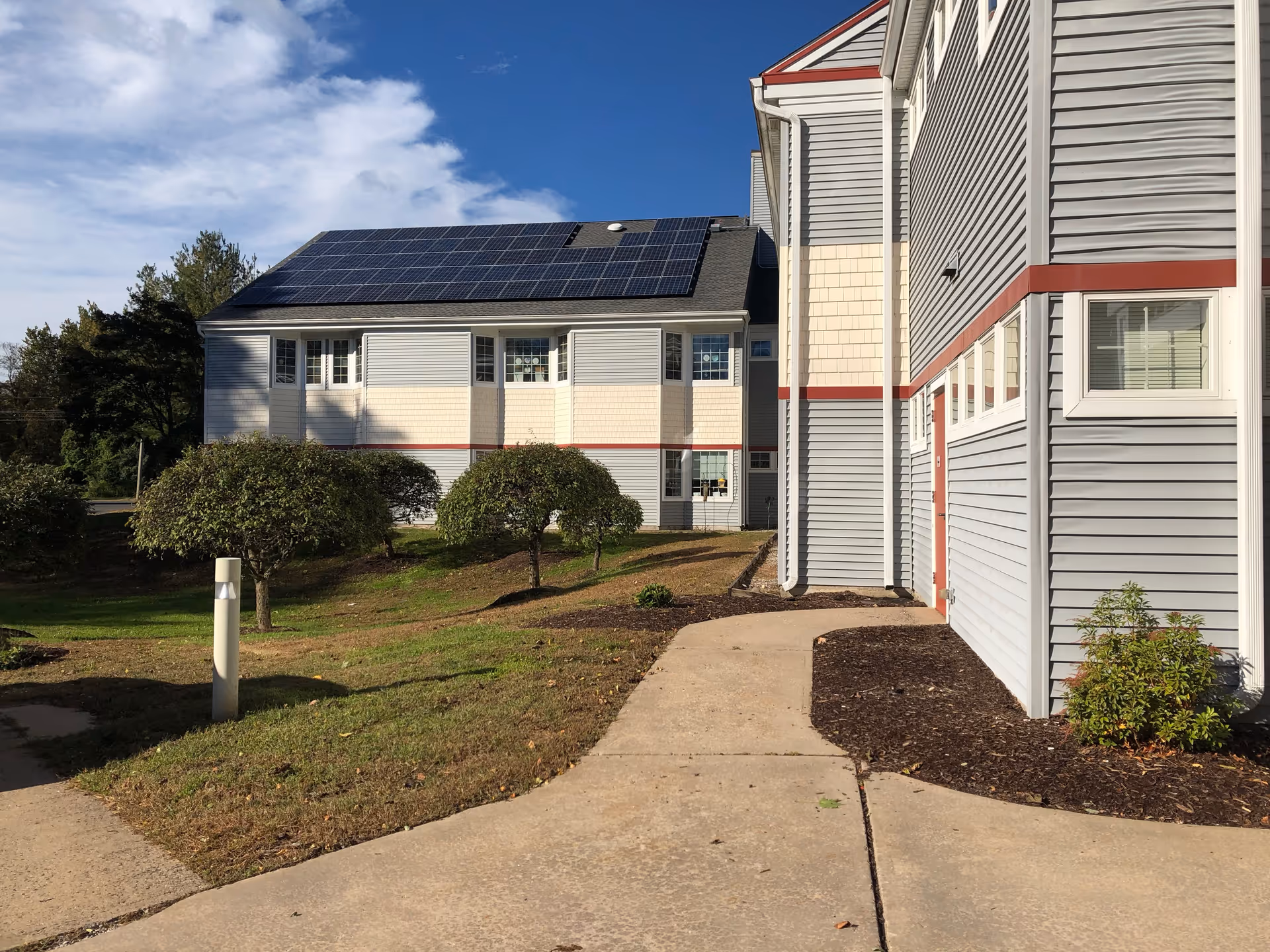 Exterior view of a senior living facility building with gray and beige siding, red trim, and solar panels on the roof. There is a concrete walkway leading alongside the building, small trimmed trees, and a grassy area under a partly cloudy blue sky.