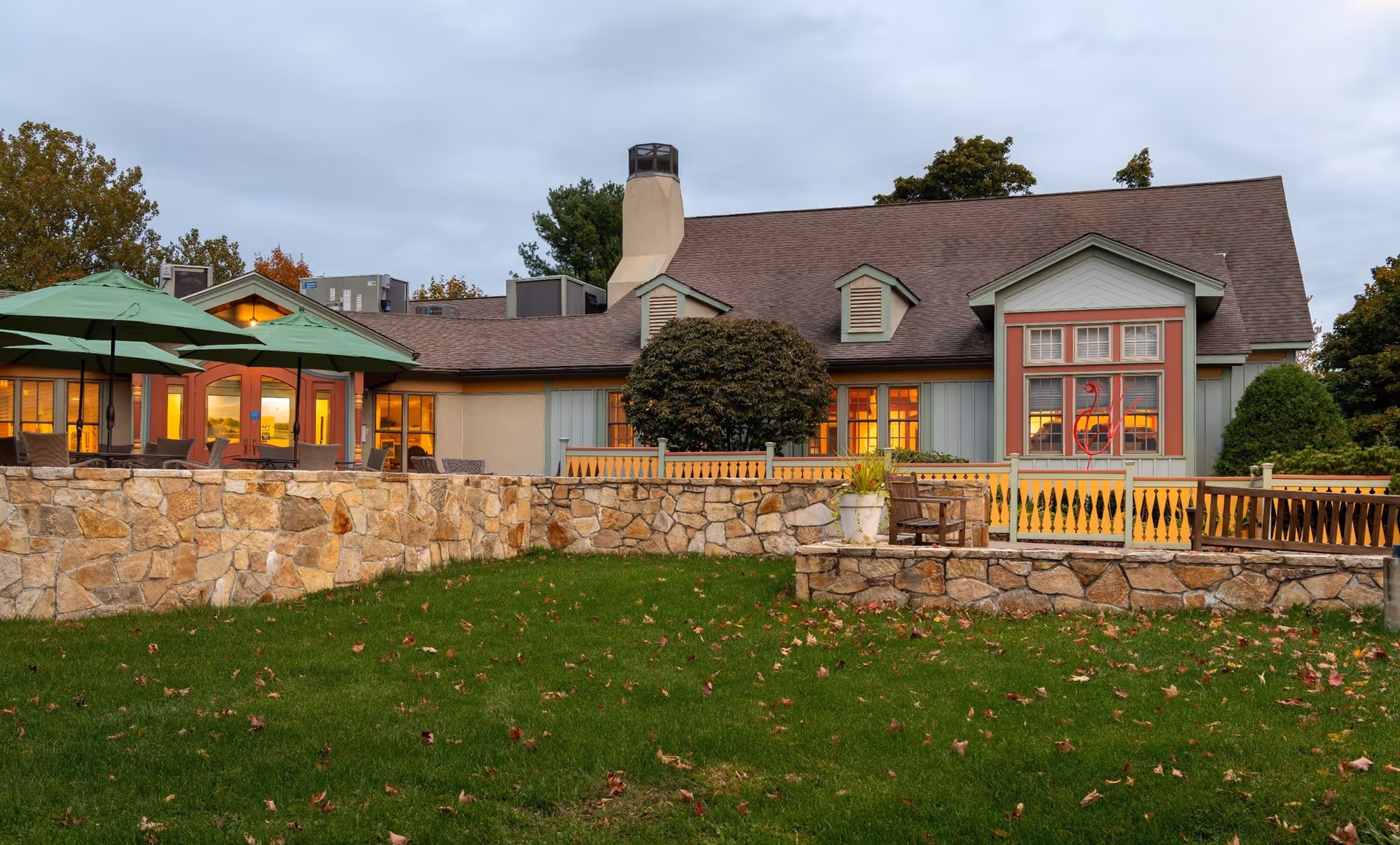 Exterior view of a single-story building with a brown shingled roof and light blue and beige walls, featuring multiple windows with warm yellow light shining through. In front of the building is a stone wall enclosing a patio area with green umbrellas and outdoor seating. The foreground shows a grassy lawn with scattered autumn leaves under a cloudy sky.