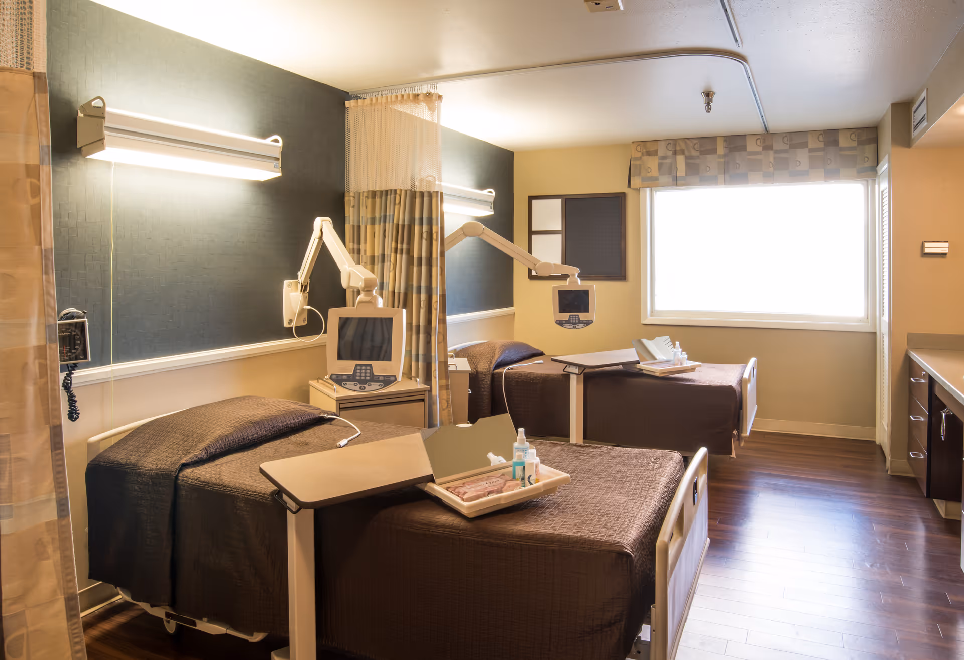 A hospital-style room with two beds separated by a privacy curtain. Each bed has a brown blanket and an adjustable overbed table with medical supplies. There are medical monitors mounted on adjustable arms above each bed. The room has a large window with a patterned valance, wood flooring, and soft lighting.