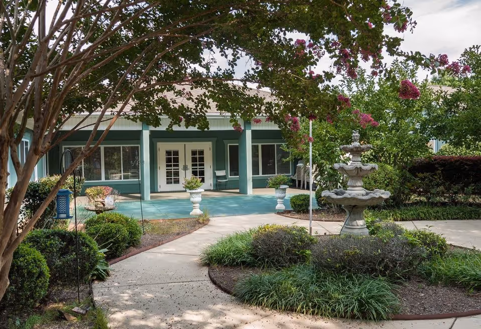 Outdoor garden area with a concrete pathway leading to a building entrance with double glass doors. The garden features green bushes, flowering trees with pink blossoms, and a tiered stone fountain. The building has green pillars and windows with white frames.