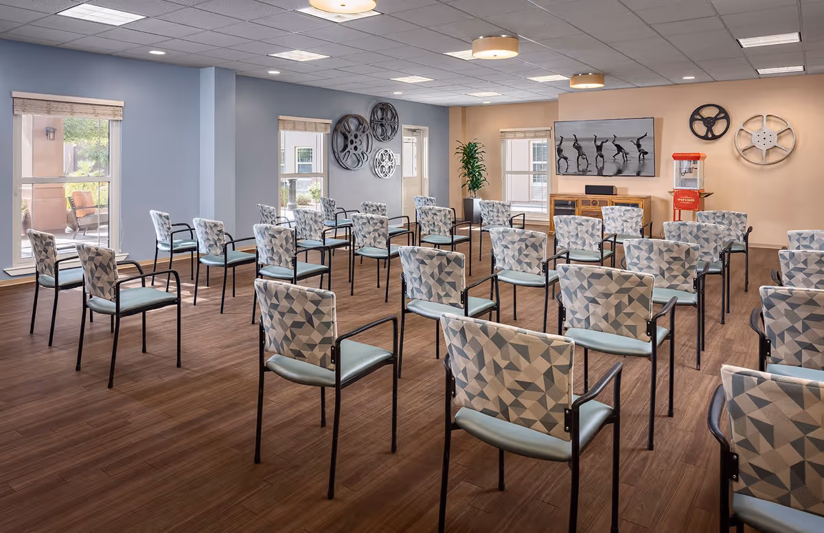 A spacious meeting or activity room with multiple rows of patterned chairs arranged facing a wall-mounted TV. The room has large windows letting in natural light, decorative film reels on the walls, a popcorn machine, and a wooden floor.