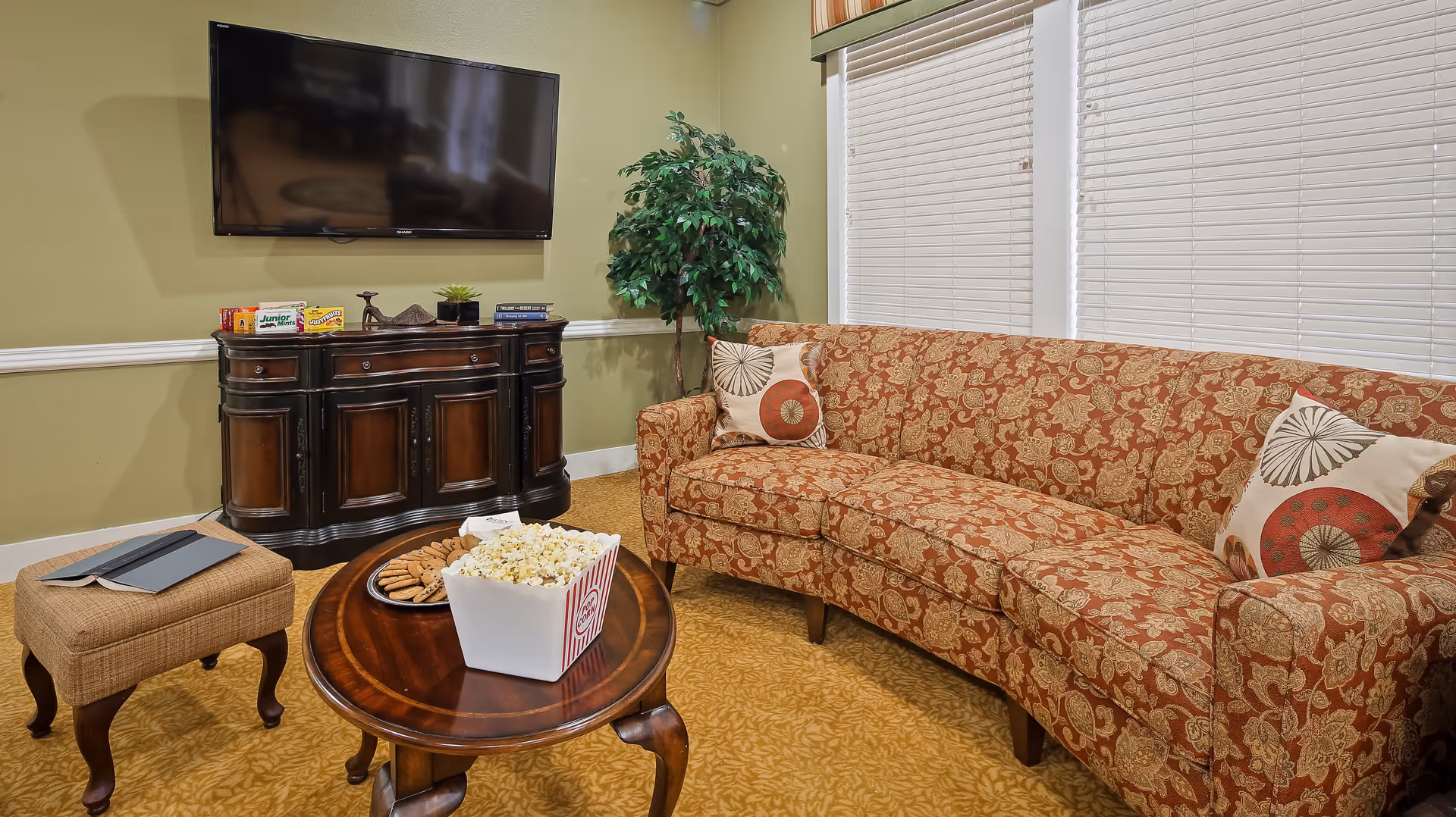 A cozy living room with a patterned orange sofa adorned with two decorative pillows, a wooden coffee table holding a container of popcorn and a plate of cookies, a small cushioned ottoman with a tablet and book on it, a dark wooden cabinet with snacks and books on top, a mounted flat-screen TV, and a potted plant in the corner. The room has light green walls and two windows with white blinds.