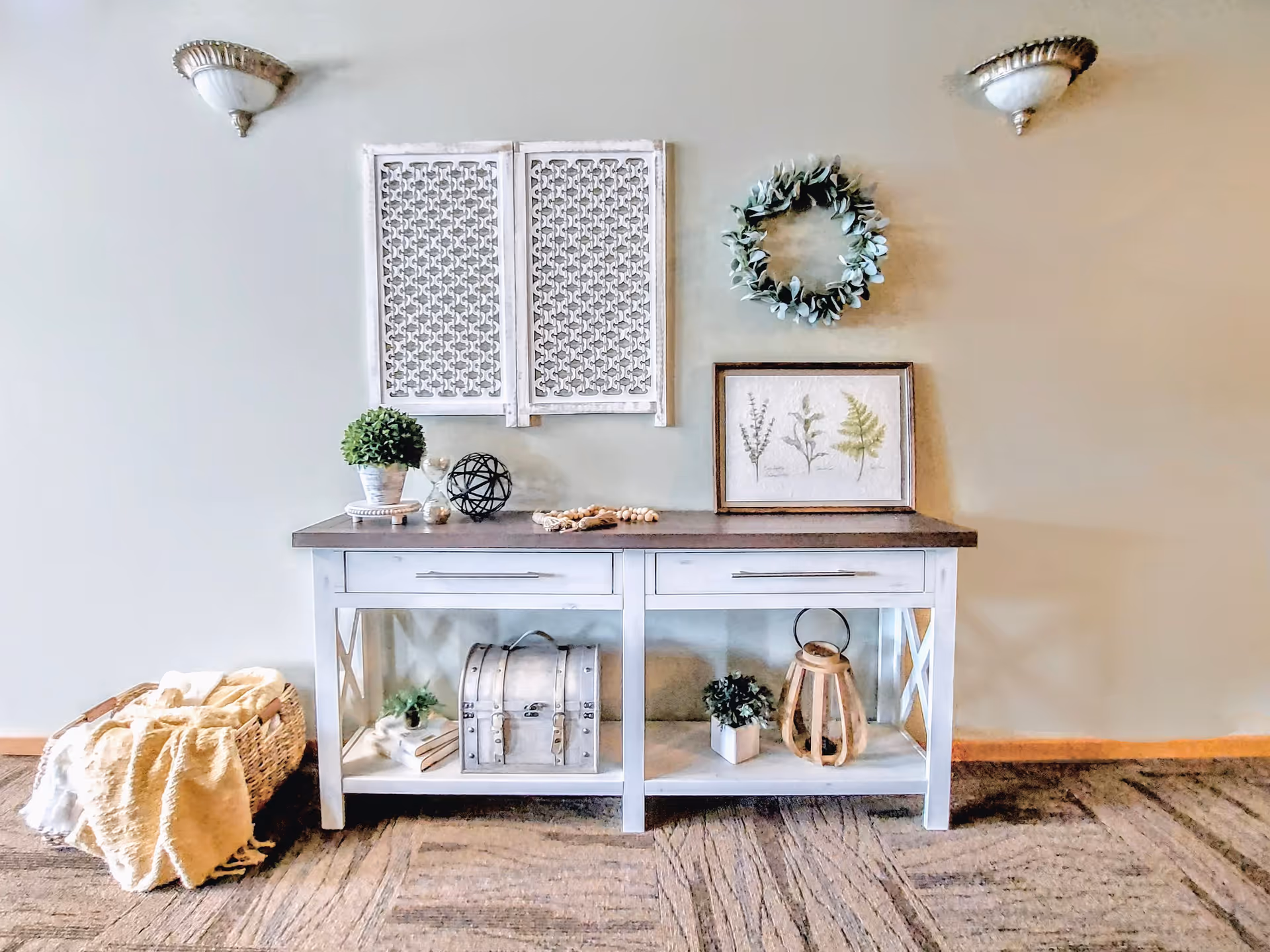A decorative console table against a beige wall with various decor items including a potted plant, an hourglass, a black decorative sphere, a wooden bead garland, a framed botanical print, a wreath, a small chest, books, a small plant, and a wooden lantern. A basket with folded blankets is placed on the floor to the left of the table. Two wall sconces are mounted above the table.