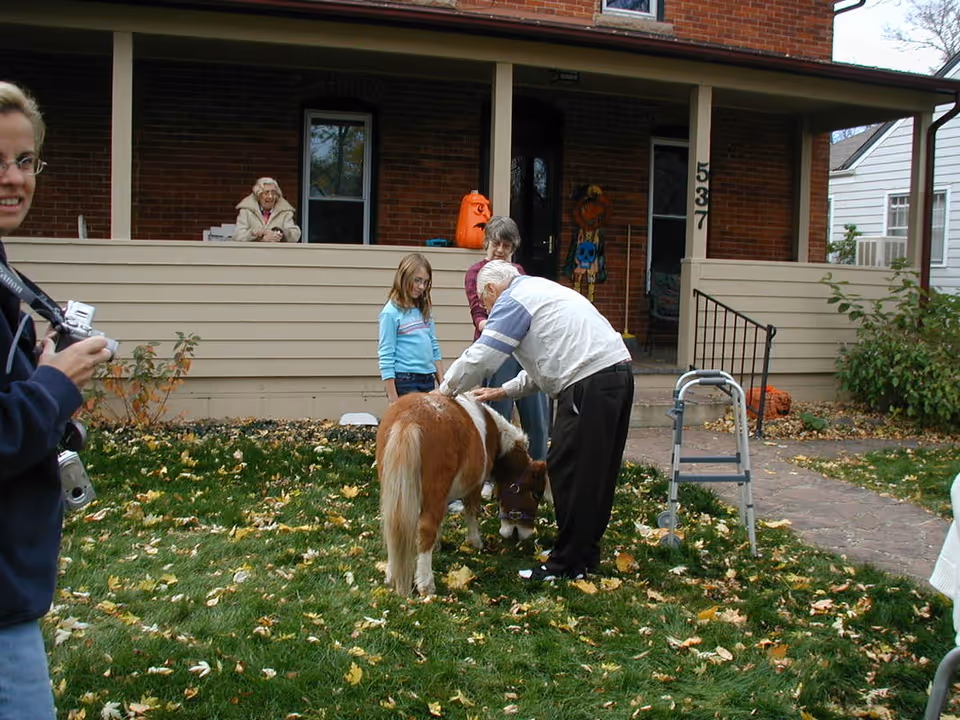 Several people, including an elderly man with a walker, a young girl, and two women, are gathered outside a brick building with a porch. The elderly man is petting a small brown and white pony on a grassy area covered with fallen leaves. Halloween decorations, including a pumpkin and a scarecrow, are visible on the porch.