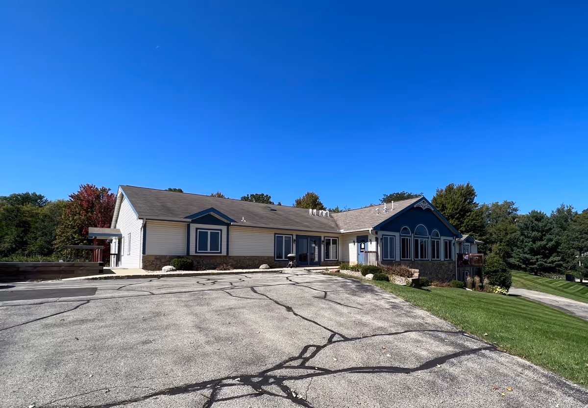 Single-story senior living building with large windows, a cracked parking lot in front, and a grassy lawn under a clear blue sky.