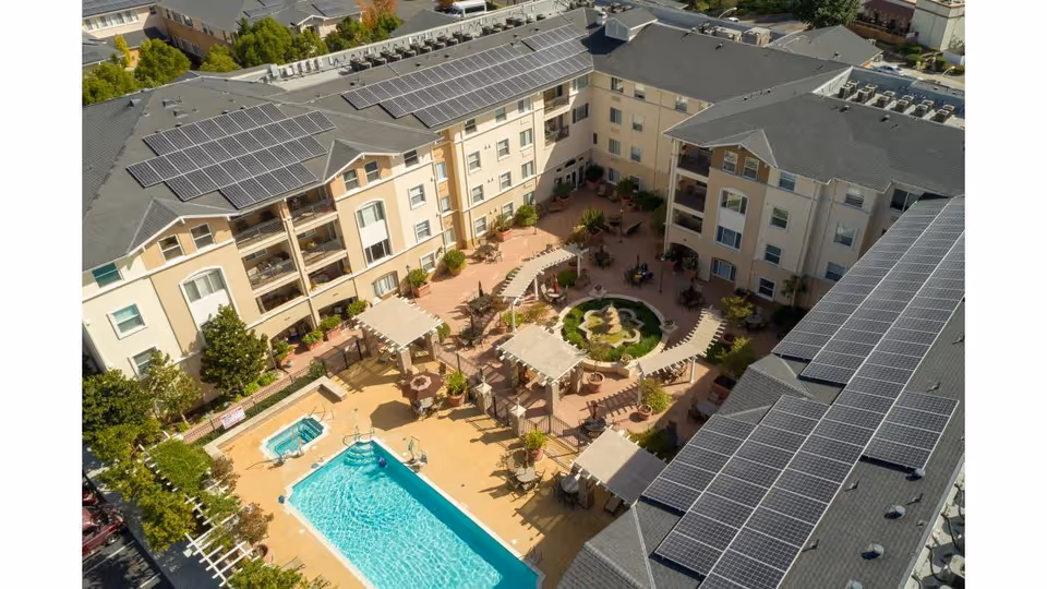 Aerial view of Heritage Estate Senior Apartments showing a large rectangular swimming pool with a smaller hot tub adjacent to it, surrounded by lounge chairs and shaded seating areas. The multi-story apartment buildings with solar panels on the roofs form a U-shape around a central courtyard featuring a circular garden with a fountain and additional seating areas.