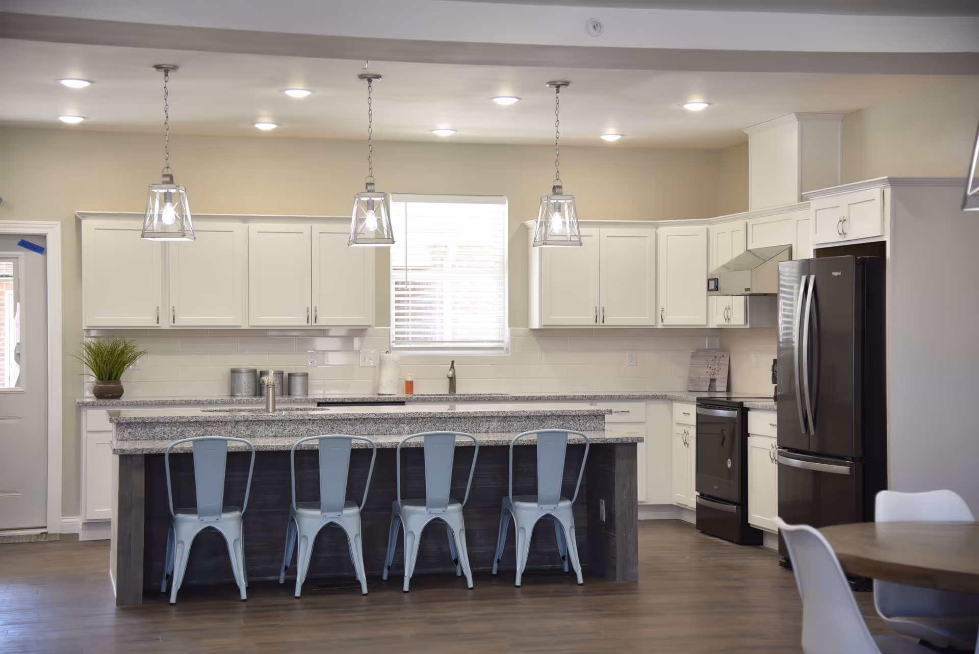 Modern kitchen with white cabinets, granite countertops, and a large island with four metal chairs. Three pendant lights hang above the island. Stainless steel appliances including a refrigerator and stove are visible. A window with blinds is centered above the sink.