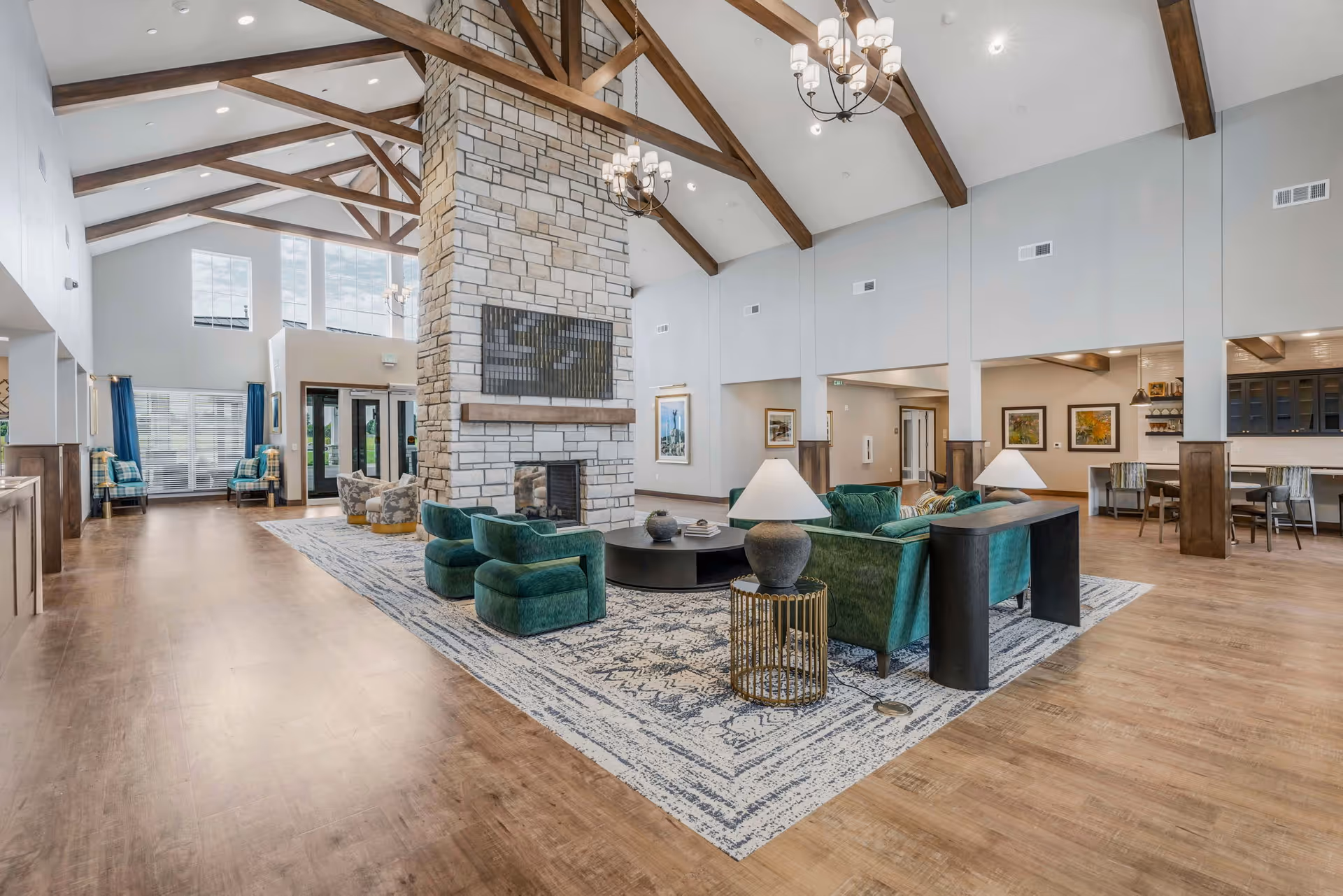 Spacious senior living facility common area with high vaulted ceilings featuring exposed wooden beams, a large stone fireplace in the center, green upholstered chairs and sofas arranged around a round coffee table on a patterned rug, and a dining area with tables and chairs in the background.