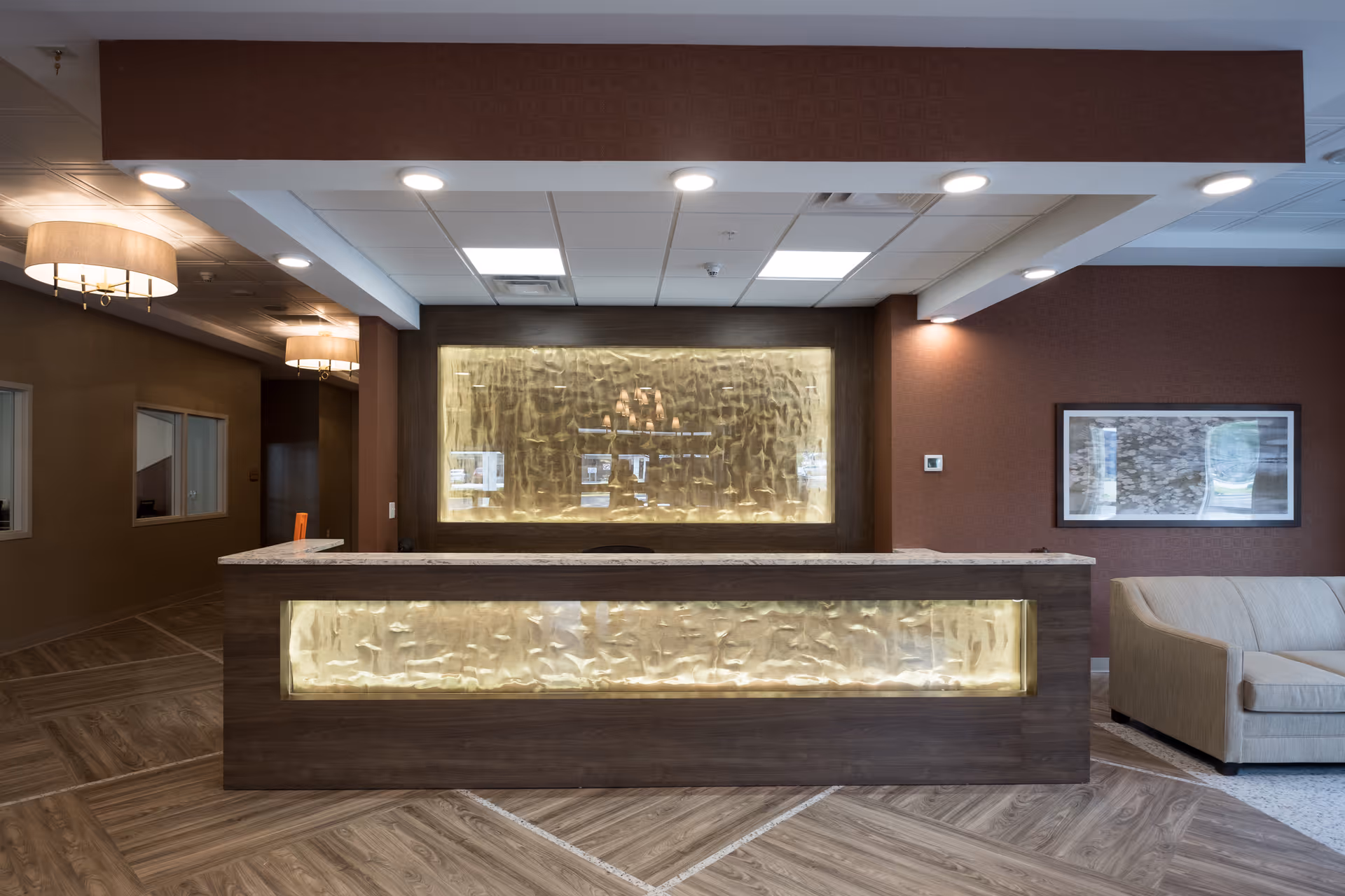 Reception area of The Sentinel of Amsterdam featuring a modern wooden front desk with illuminated textured panels, overhead recessed lighting, two wall-mounted lamps, a beige couch to the right, and a framed picture on the wall.