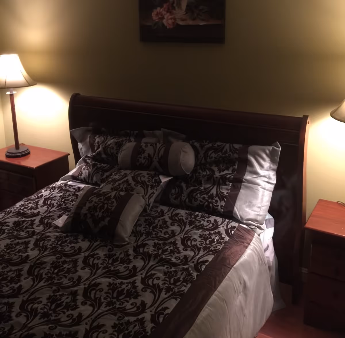 A bedroom with a wooden bed frame and a bed made with patterned bedding in shades of brown and beige. There are two matching wooden nightstands on either side of the bed, each with a lit table lamp. A floral painting hangs on the wall above the bed.