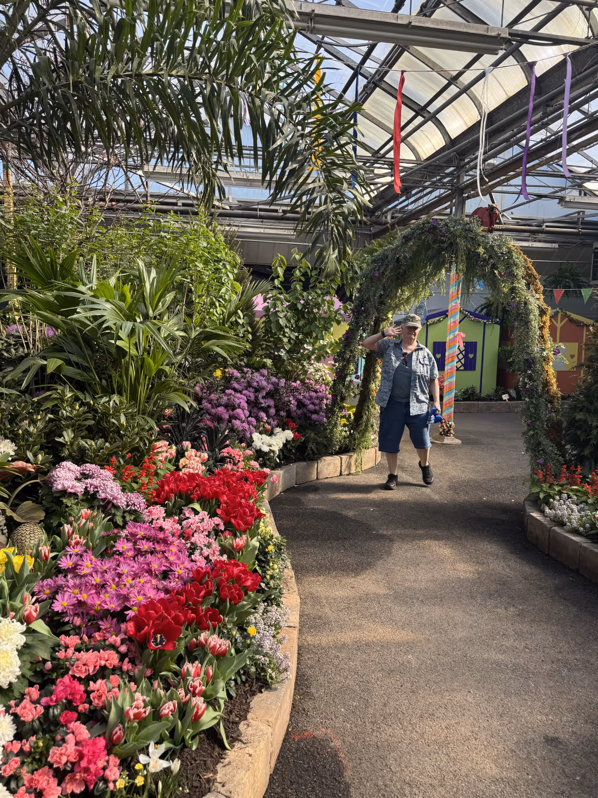 A greenhouse walkway filled with colorful flower beds and plants, with a person walking under a leafy decorative arch.
