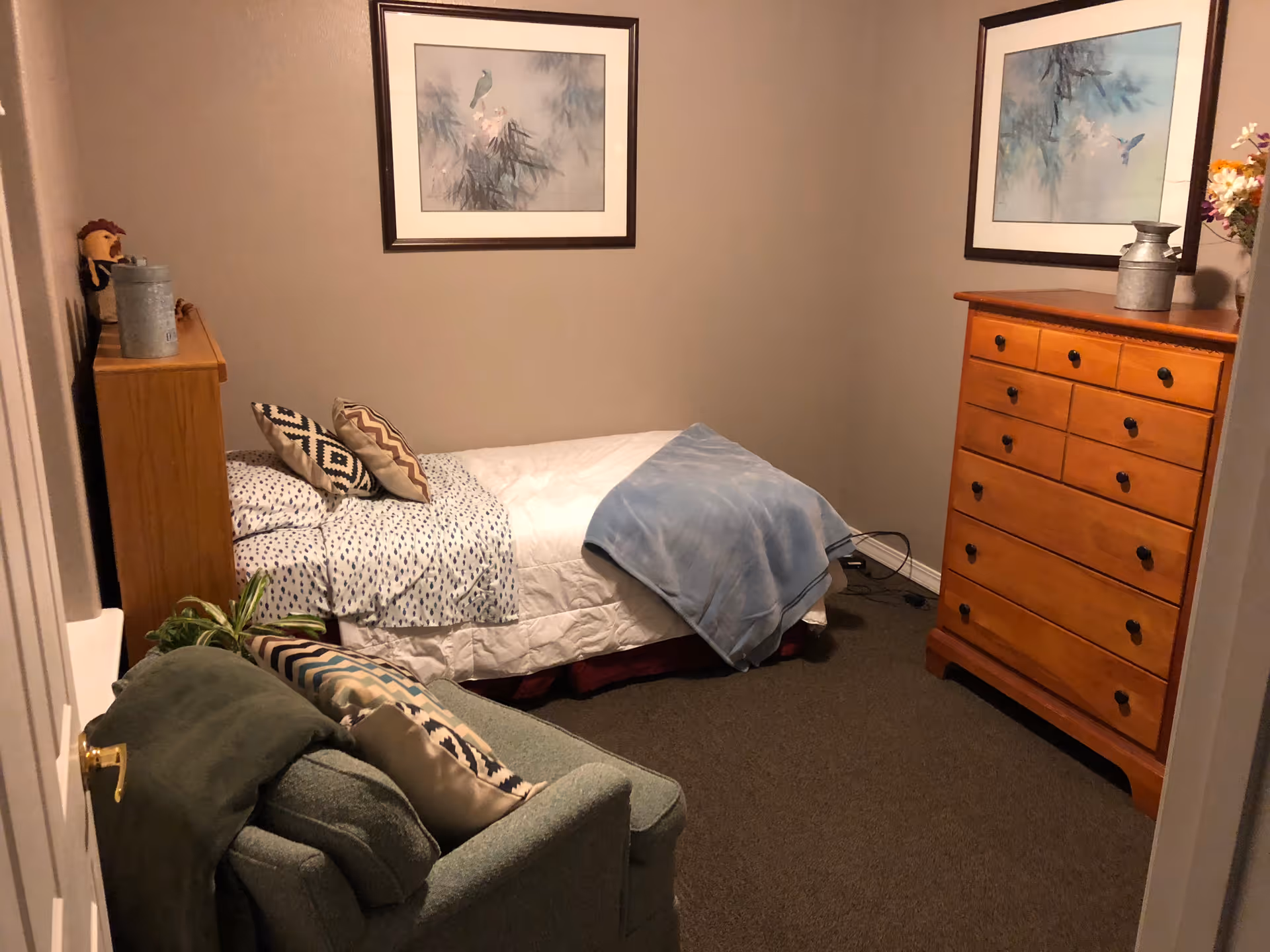 A small bedroom with a single bed covered with white and blue bedding and two patterned pillows. There is a wooden dresser with multiple drawers on the right side and a small wooden shelf at the foot of the bed with decorative items. A green armchair with patterned cushions is positioned near the door. Two framed pictures of birds hang on the beige walls.